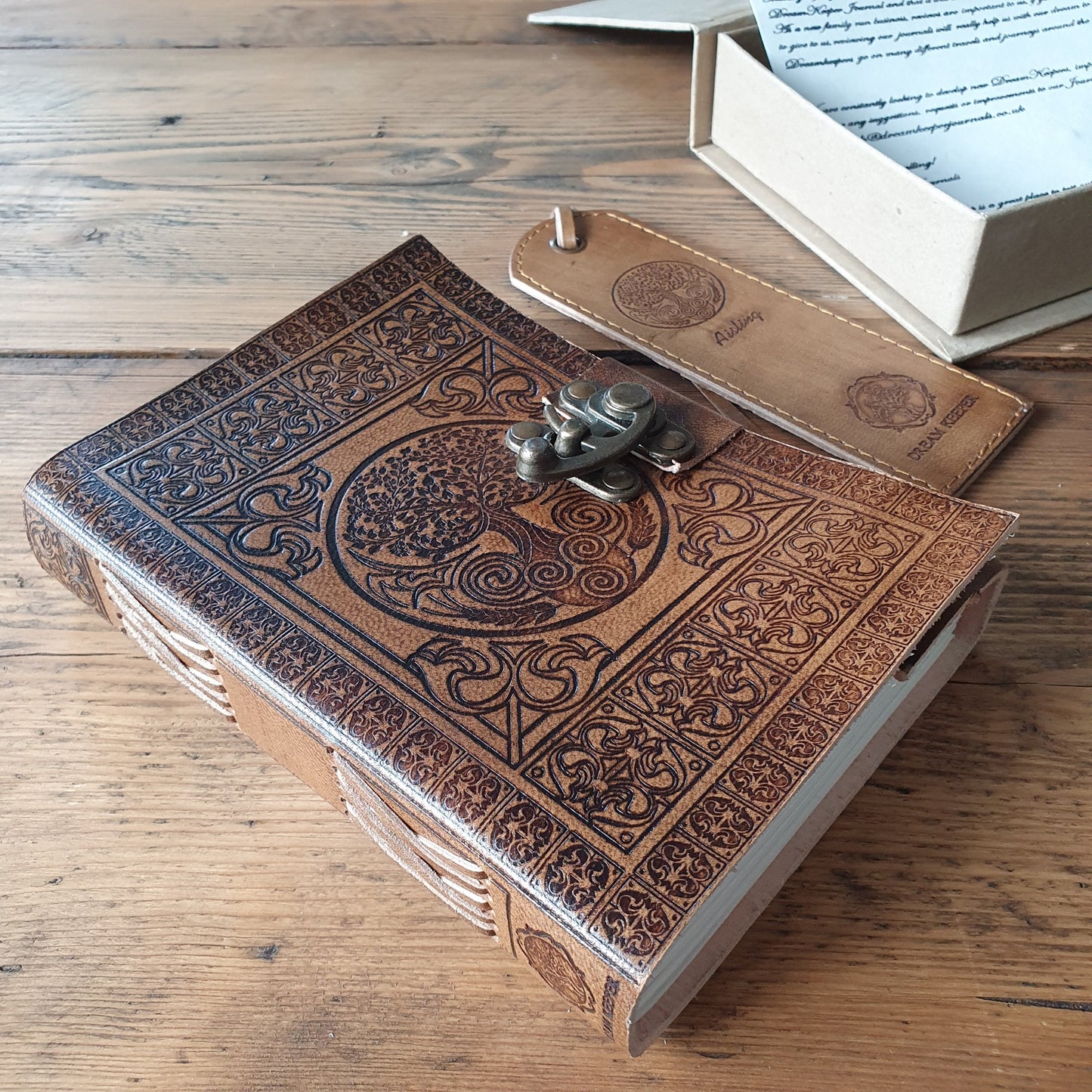 Tree of Life embossed notebook resting on wooden table beside leather bookmark and protective delivery box
