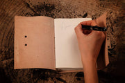 Leather journal open, revealing recycled white pages with a hand scribbling words